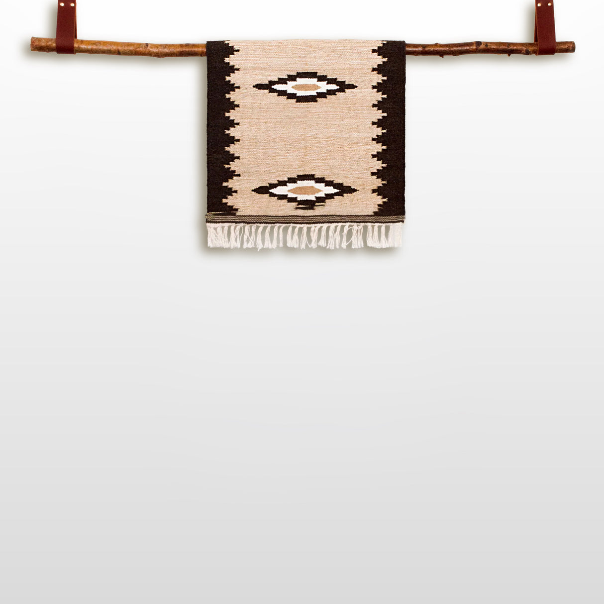 The Heredij (Heritage) Wool Rug, a handmade beige and black geometric wall hanging crafted from natural wool, is displayed on a wooden branch against a white background.