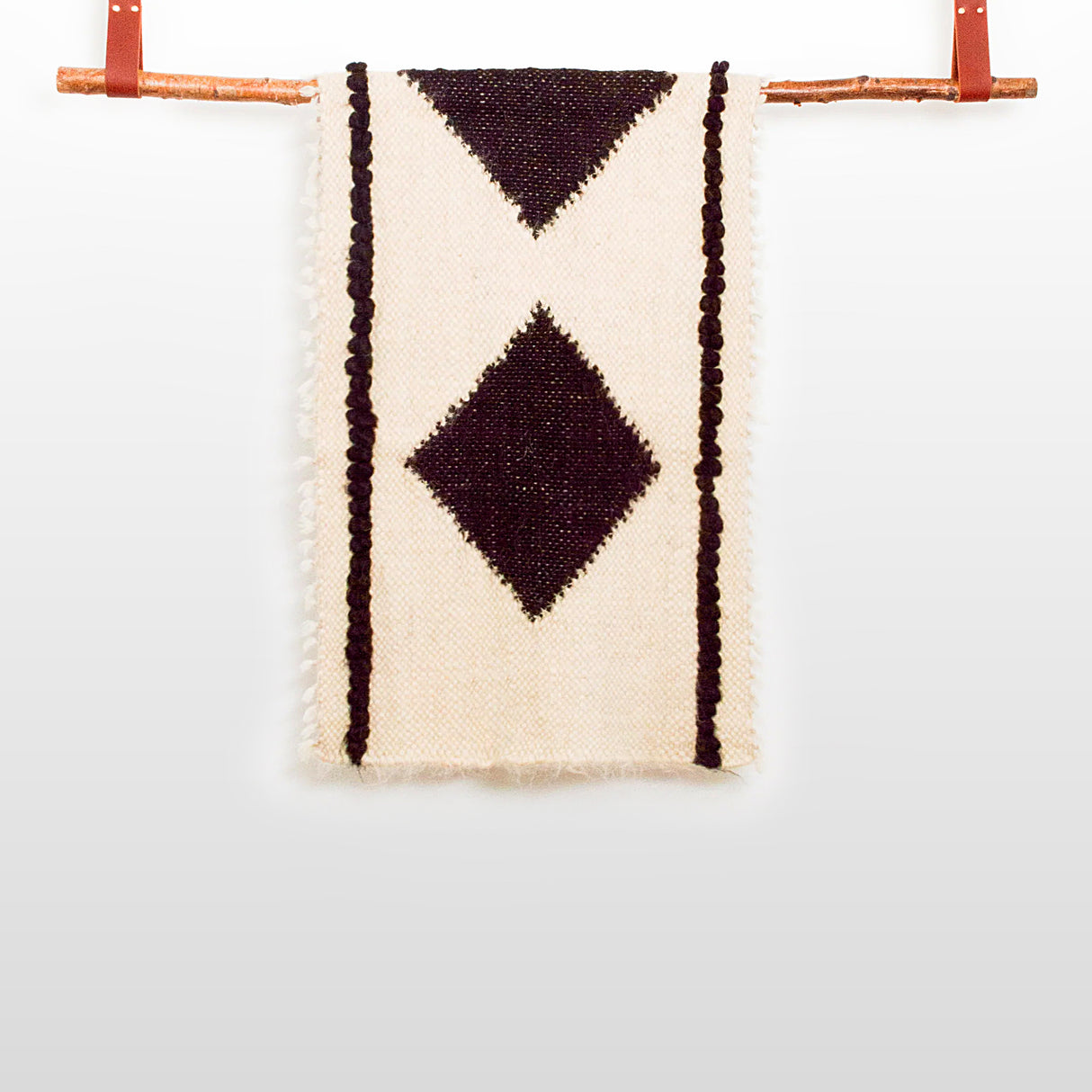 The Korysno (Helpful) Wool Bed/Couch Runner is handcrafted from 100% virgin wool in beige, with black diamond and stripe patterns, and is displayed hanging on a wooden rod against a white background.