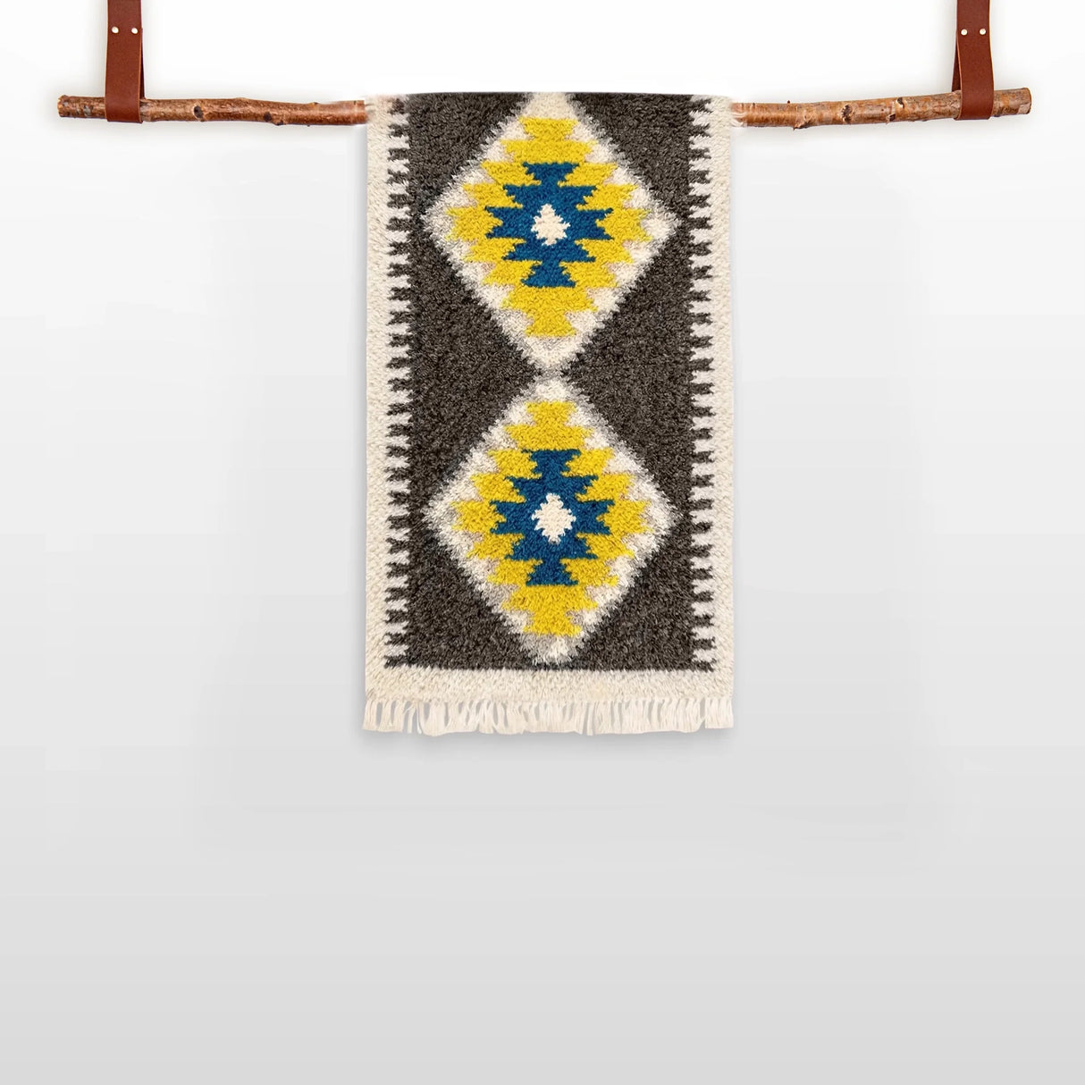 The Mriya (Dream) Wool Bed / Couch Runner, featuring yellow, blue, and white diamond patterns and Ukrainian craftsmanship, hangs from a wooden rod with leather straps.