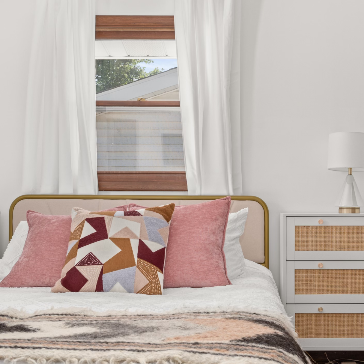 Cozy bed adorned with pink and patterned pillows, a Tkach (Weaver) Wool Statement Bed Blanket, white curtains, and a nightstand topped with a lamp.