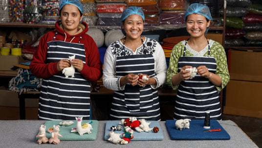 Three women in striped aprons and hairnets smile while crafting felt animal figures at a workshop table.