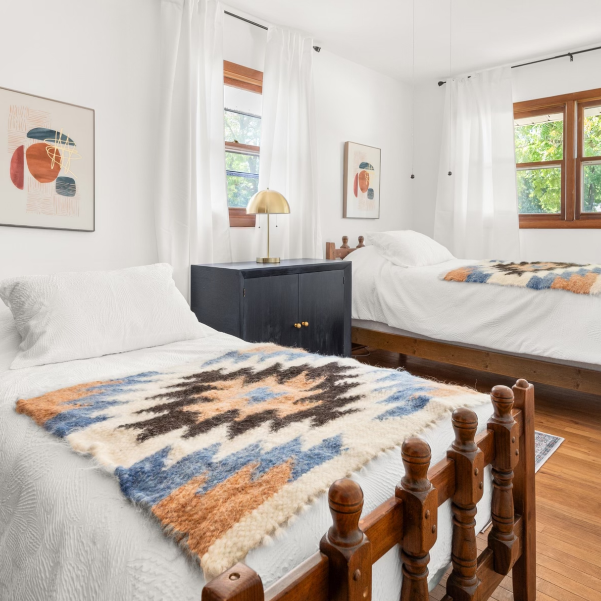 Two wooden twin beds with patterned blankets and the Zirka (Star) Wool Wall Hanging decorate a bright, white-walled bedroom with large windows.