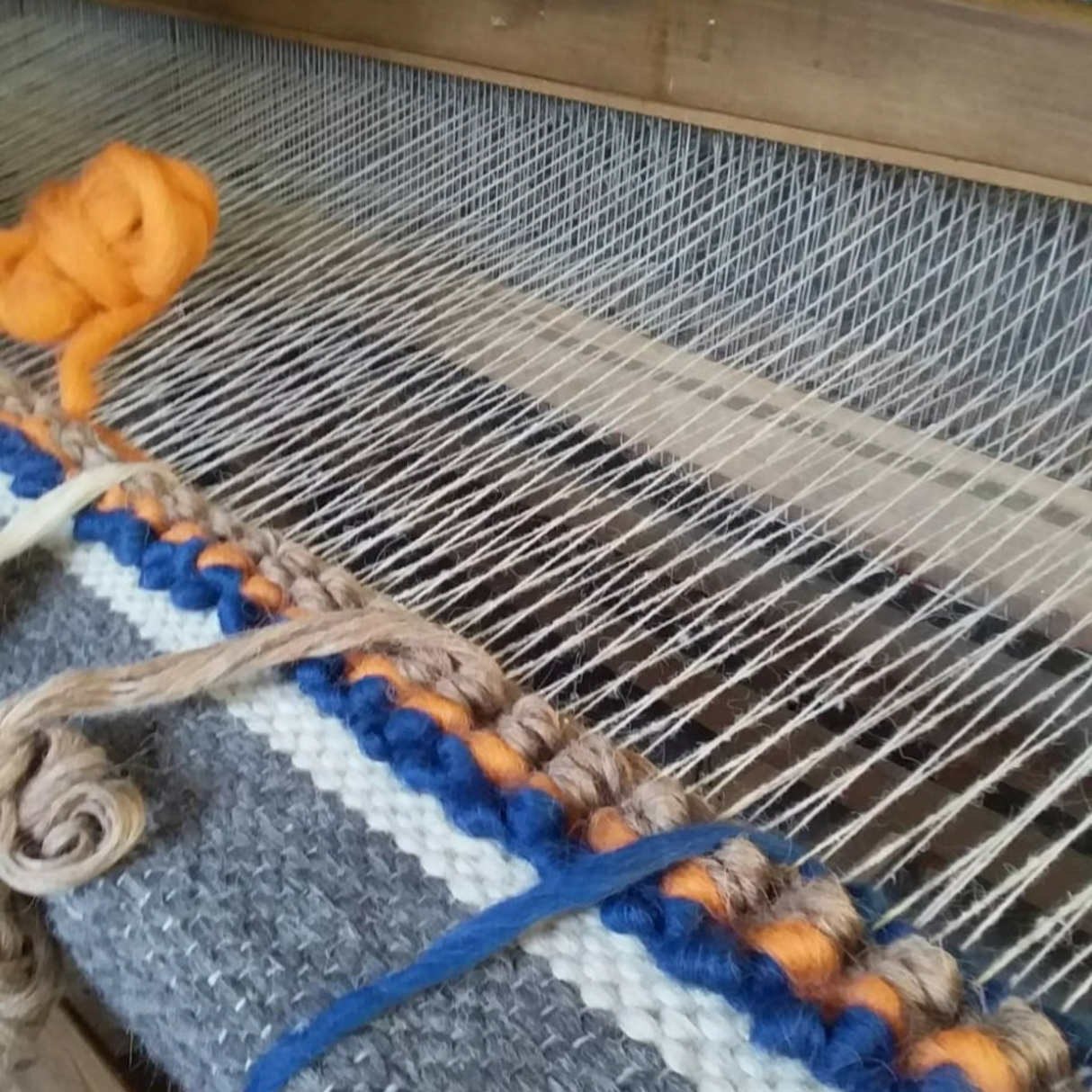 Close-up of the Tkach (Weaver) Wool Statement Bed Blanket on a loom, featuring blue, orange, white, and brown yarns being woven—an artisanal, colorful piece ideal for unique home décor.