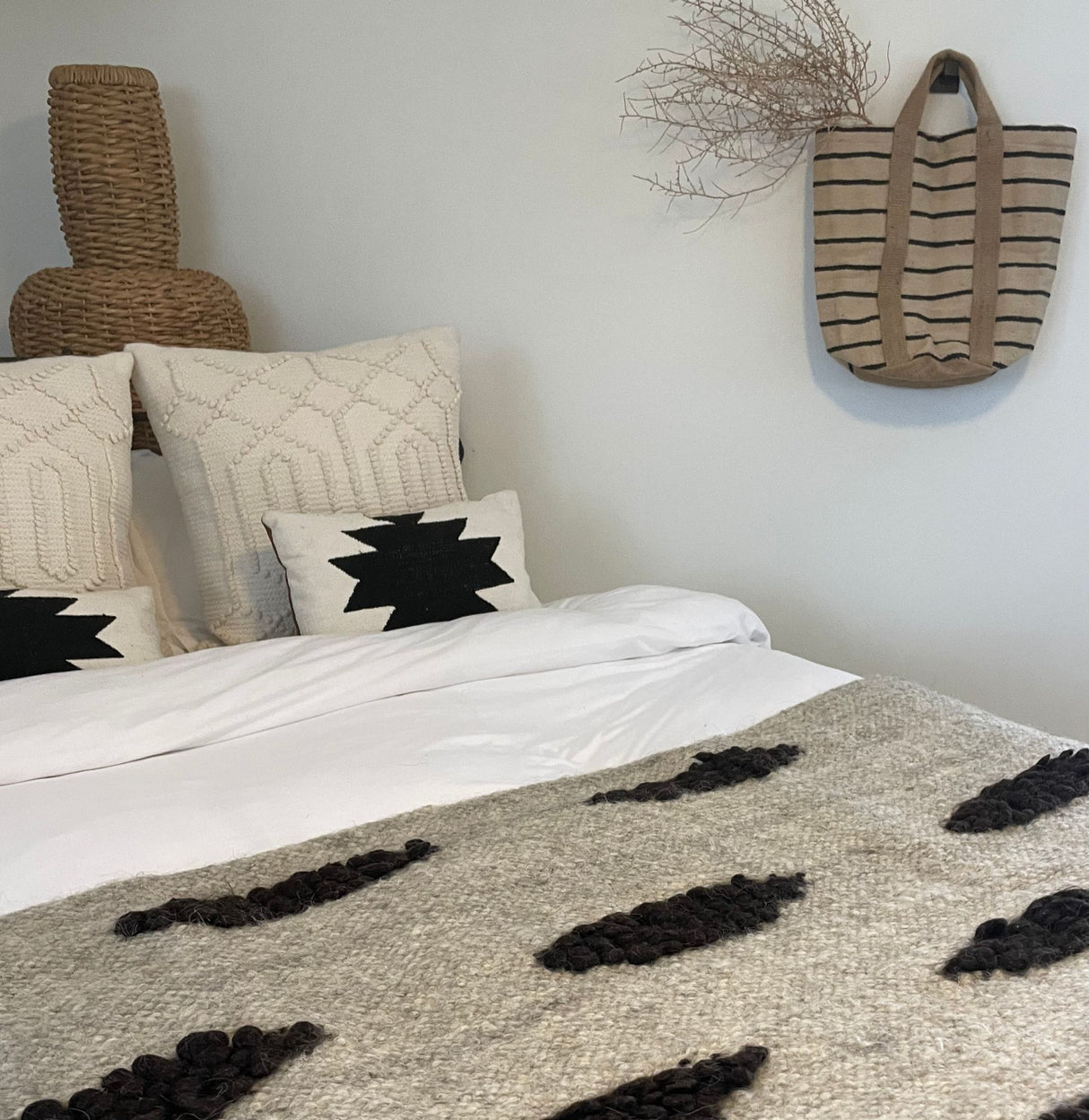 Neutral-toned bed with patterned pillows, Khmara (Cloud) Wool Statement Bed Blanket, woven basket, and a striped tote hung on the wall with dried branches.
