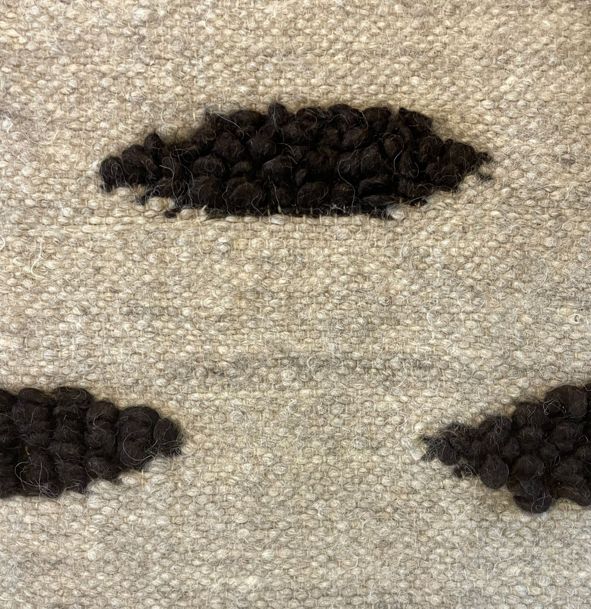 Close-up of beige woven fabric from the Khmara (Cloud) Wool Statement Bed Blanket, featuring three raised black textured patches arranged in a pattern.