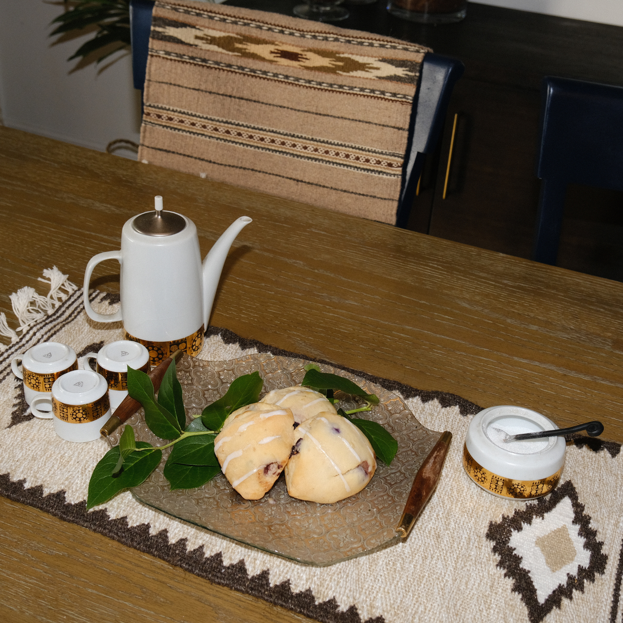 A teapot, cups, and pastries on a decorated table with green leaves and a woven runner, alongside the Heredij (Heritage) Wool Rug / Wall Hanging—handcrafted from natural wool and inspired by Carpathian mountain tradition.