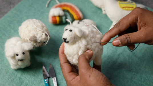 Hands needle felting a wool sheep figure, with other felted animals and scissors nearby on a green surface.