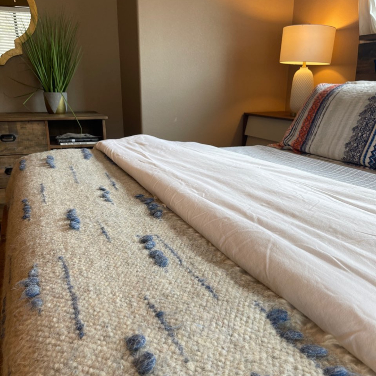 A neatly made bed with striped pillows, the Horoshy (Kind) Wool Statement Bed Blanket inspired by Ukrainian weavers in beige, and a lit lamp on a nightstand.