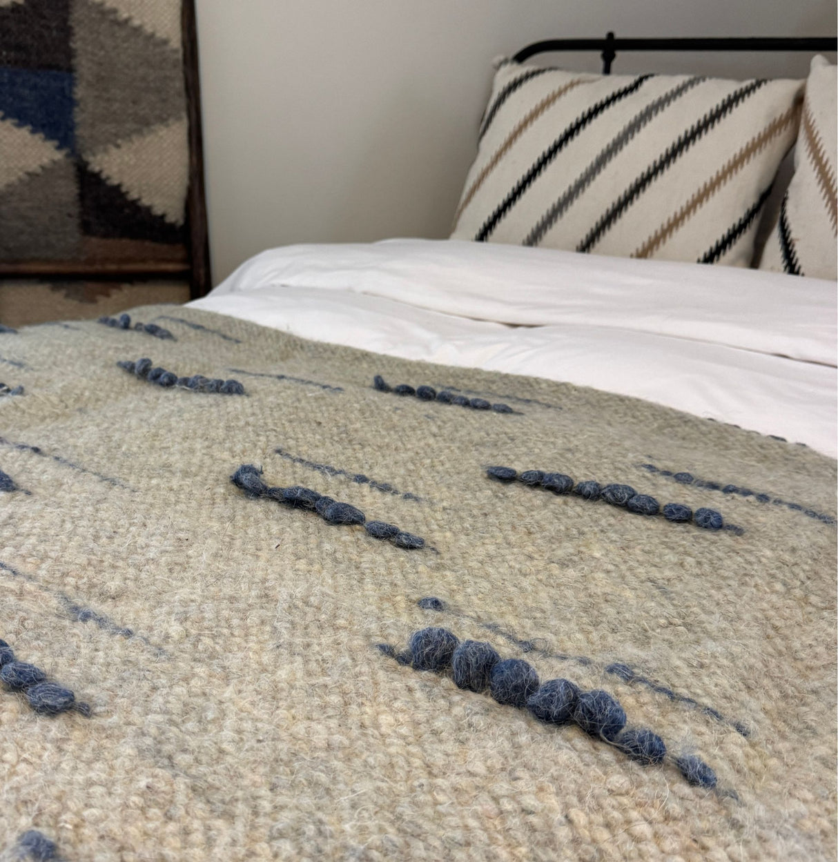 A close-up of the Horoshy (Kind) Wool Statement Bed Blanket, handwoven from organic cruelty-free wool, featuring a beige texture with blue accents, displayed on a neatly made bed with striped pillows.
