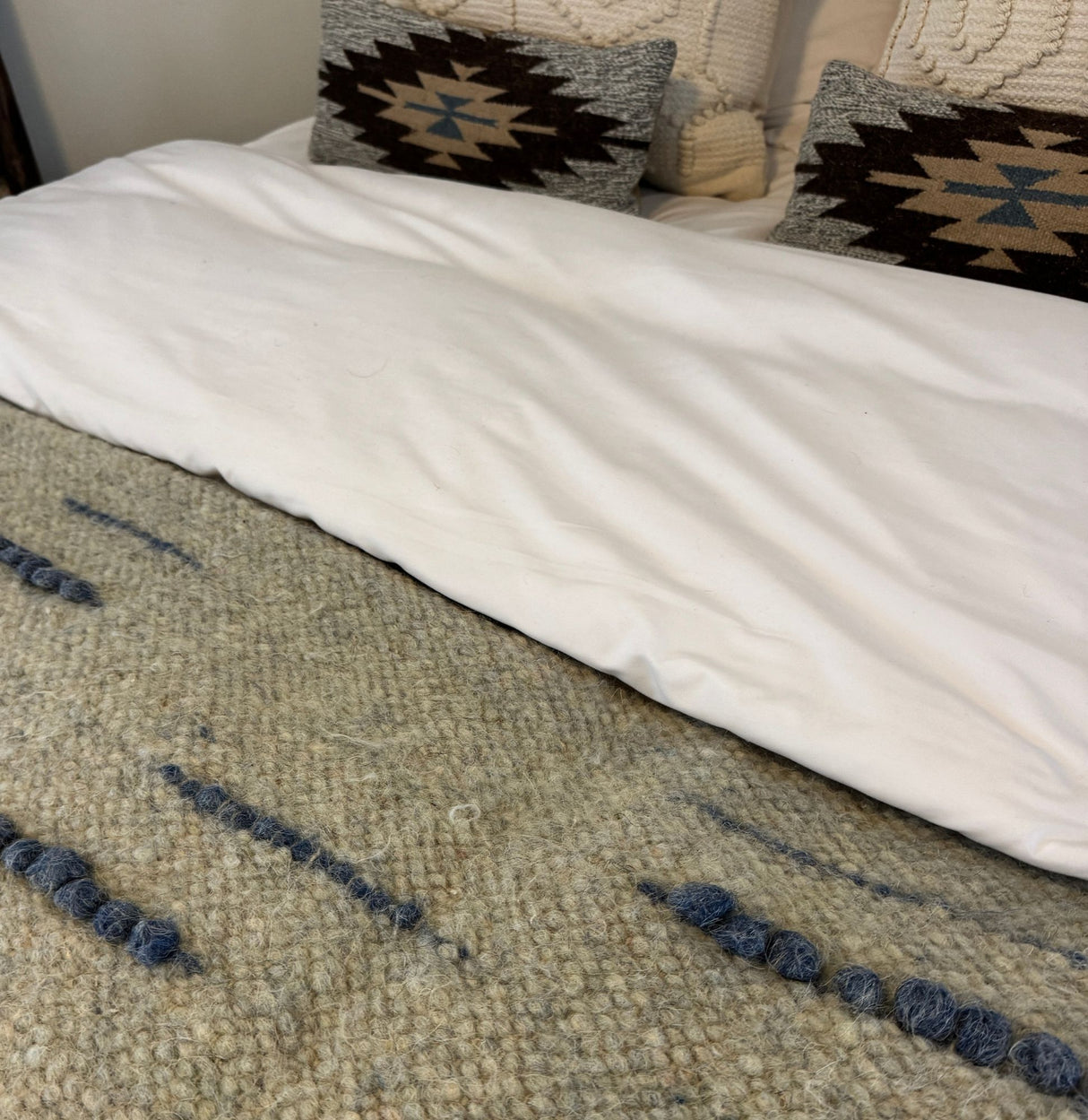 A bed with a white duvet, the Horoshy (Kind) Wool Statement Bed Blanket made from organic cruelty-free wool, and two geometric patterned pillows.