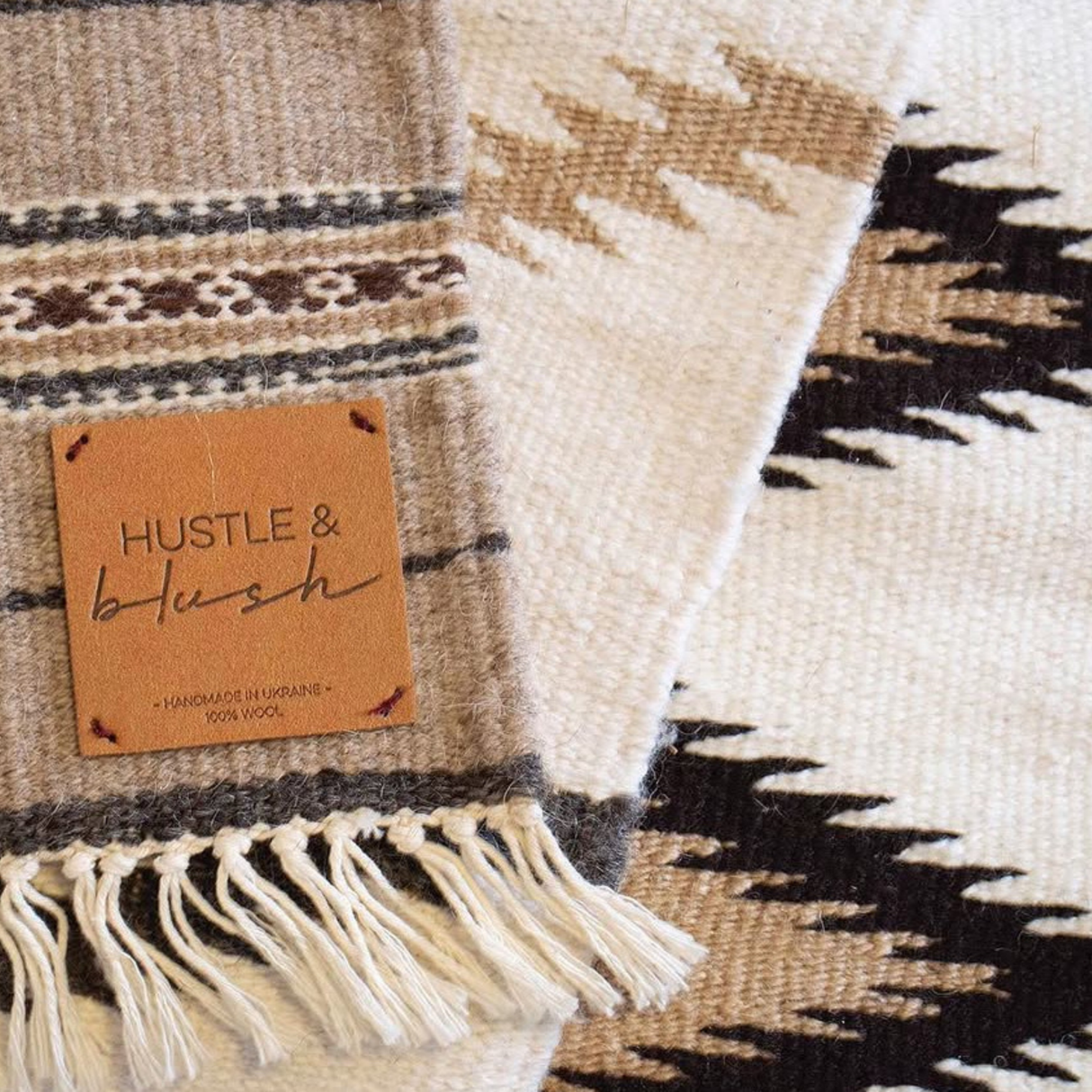 Close-up of the Promin (Beam) Wool Rug / Wall Hanging, woven from Carpathian mountain sheep wool, featuring a "Hustle & Blush" leather tag and beige, brown, and black patterns—perfect for cozying up by a gallery wall.