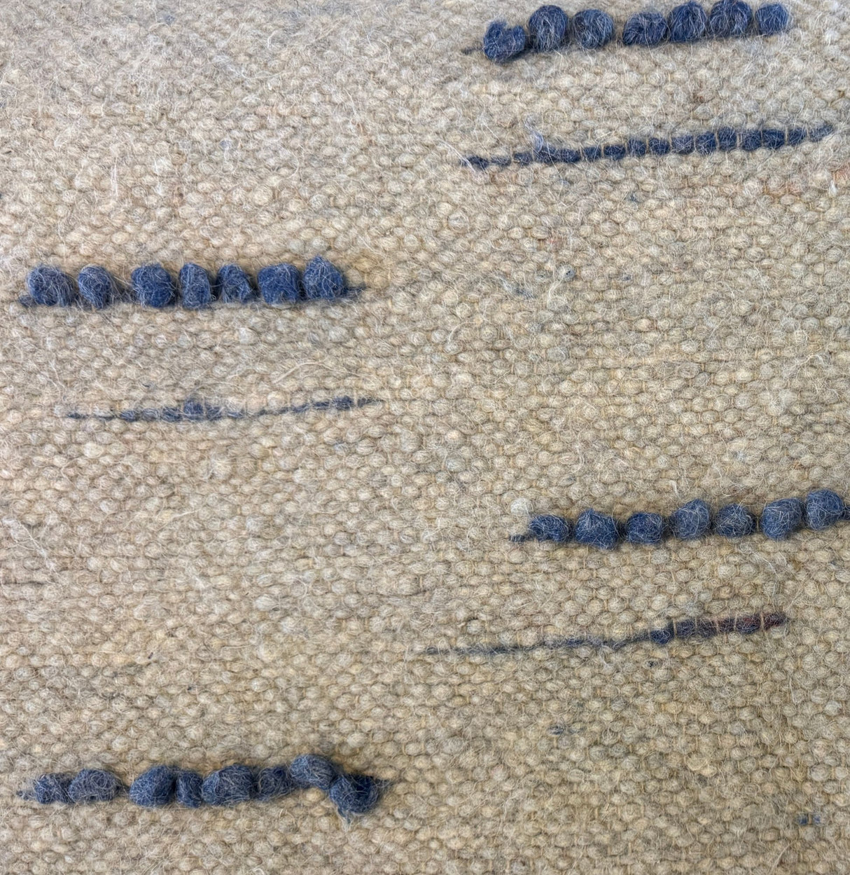 Close-up of the Horoshy (Kind) Wool Statement Bed Blanket: textured beige fabric, handwoven from organic cruelty-free wool, features small uneven rows of blue yarn stitched throughout.