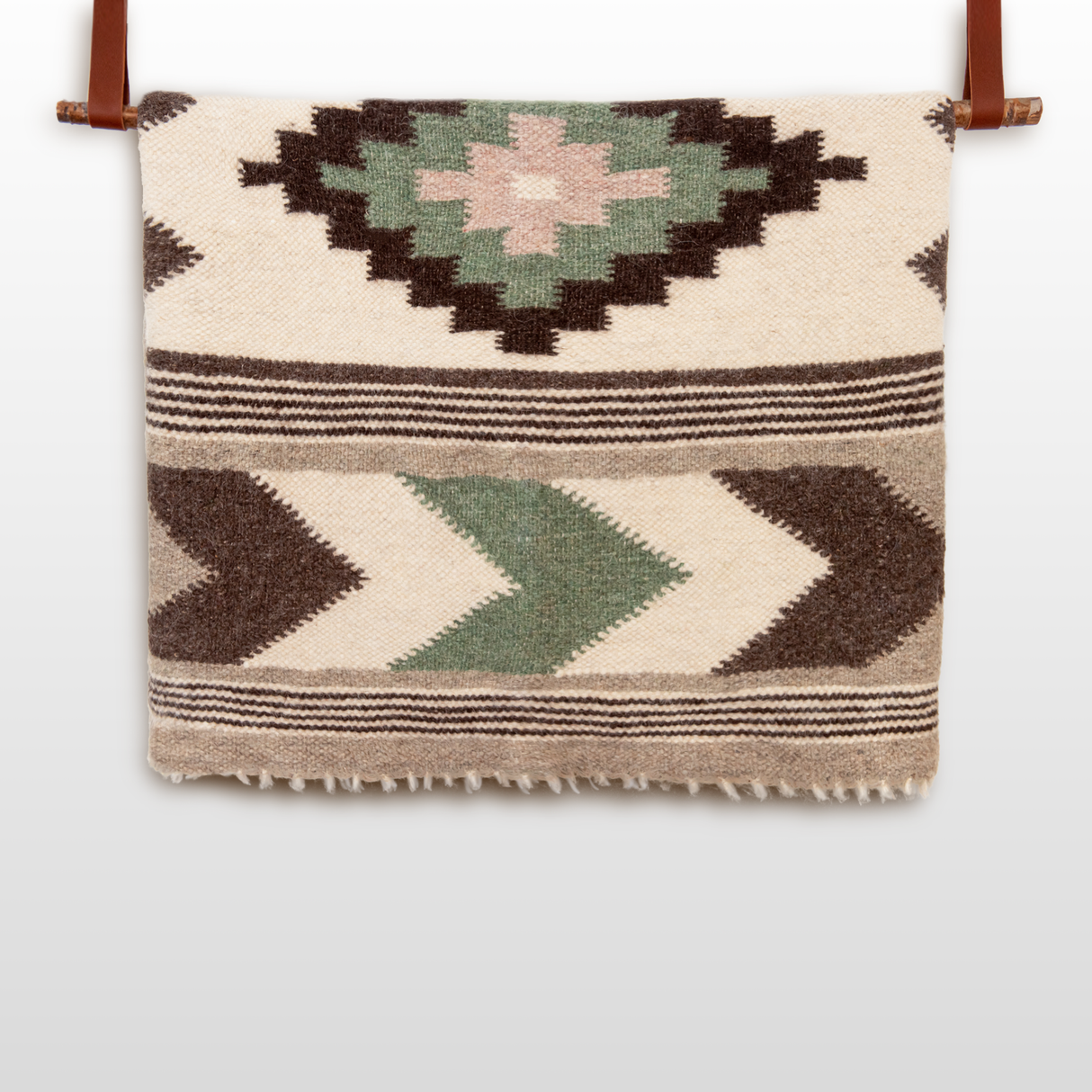 The Myr (Peace) Wool Statement Bed Blanket, featuring geometric patterns in green, brown, and beige, hangs elegantly from a leather strap.