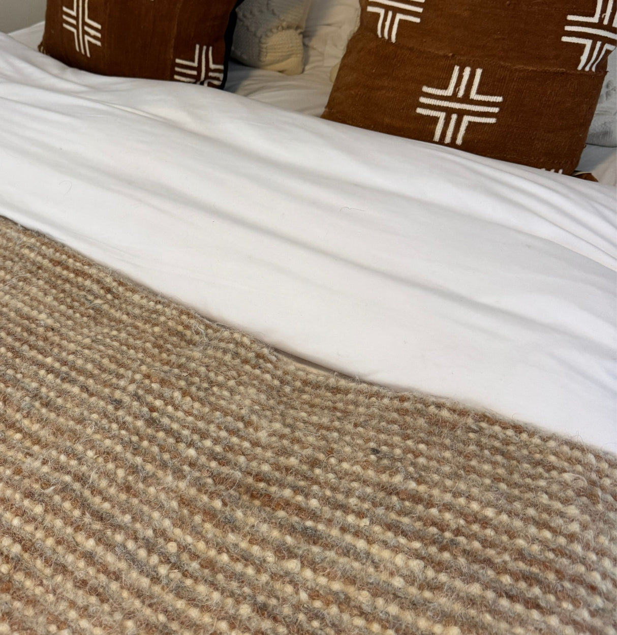 A cozy bed features a white duvet, brown patterned pillows, and the Tkach (Weaver) Wool Statement Bed Blanket, a textured beige handwoven wool accent.