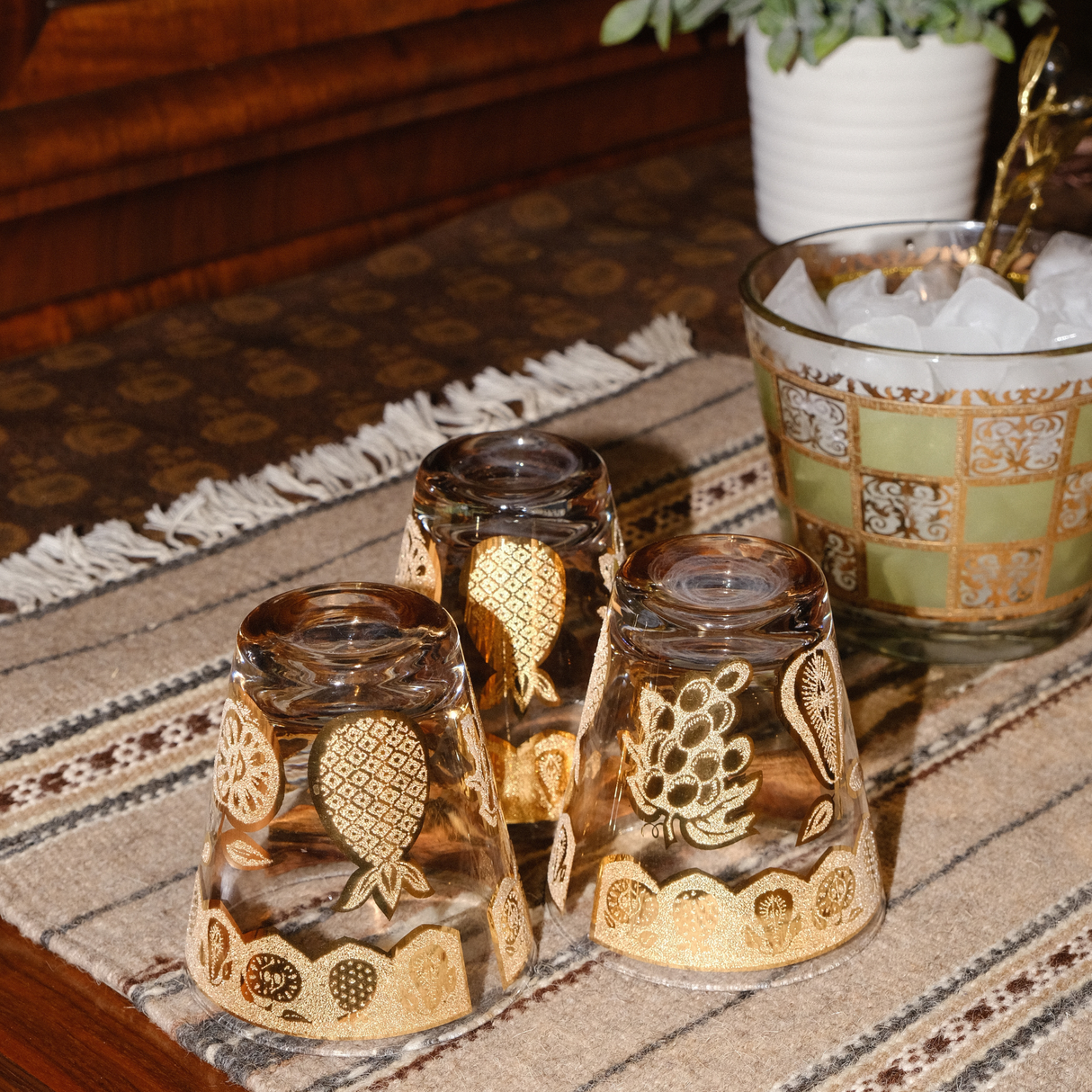 Three upside-down glasses with gold fruit designs rest on the Teplota (Warmth) Wool Small Rug / Mat, beside a glass of ice, adding a cozy touch to your decor.
