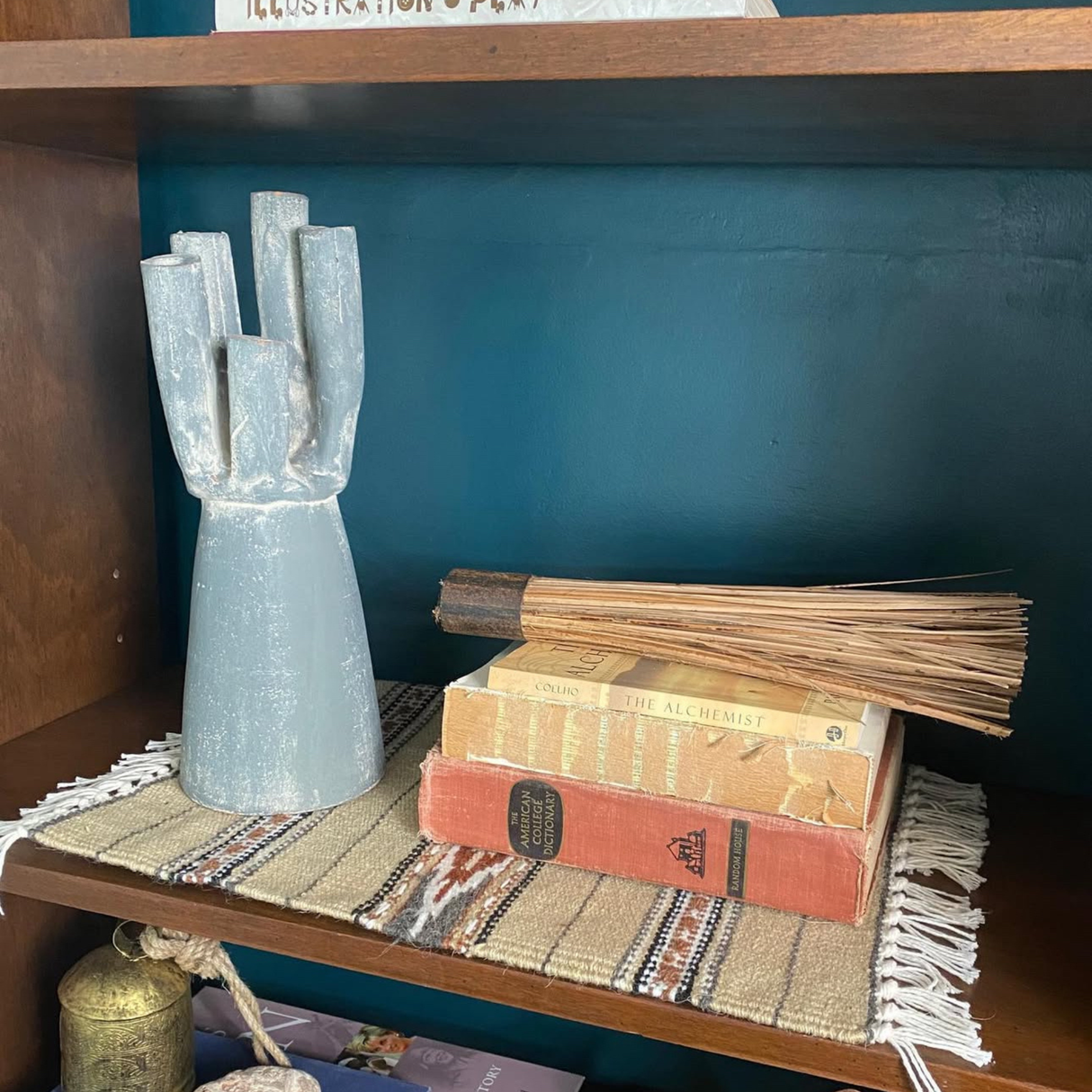 The Teplota (Warmth) Wool Small Rug / Mat sits on a wooden shelf against a blue wall, topped with a ceramic vase, stacked books, and a decorative broom.