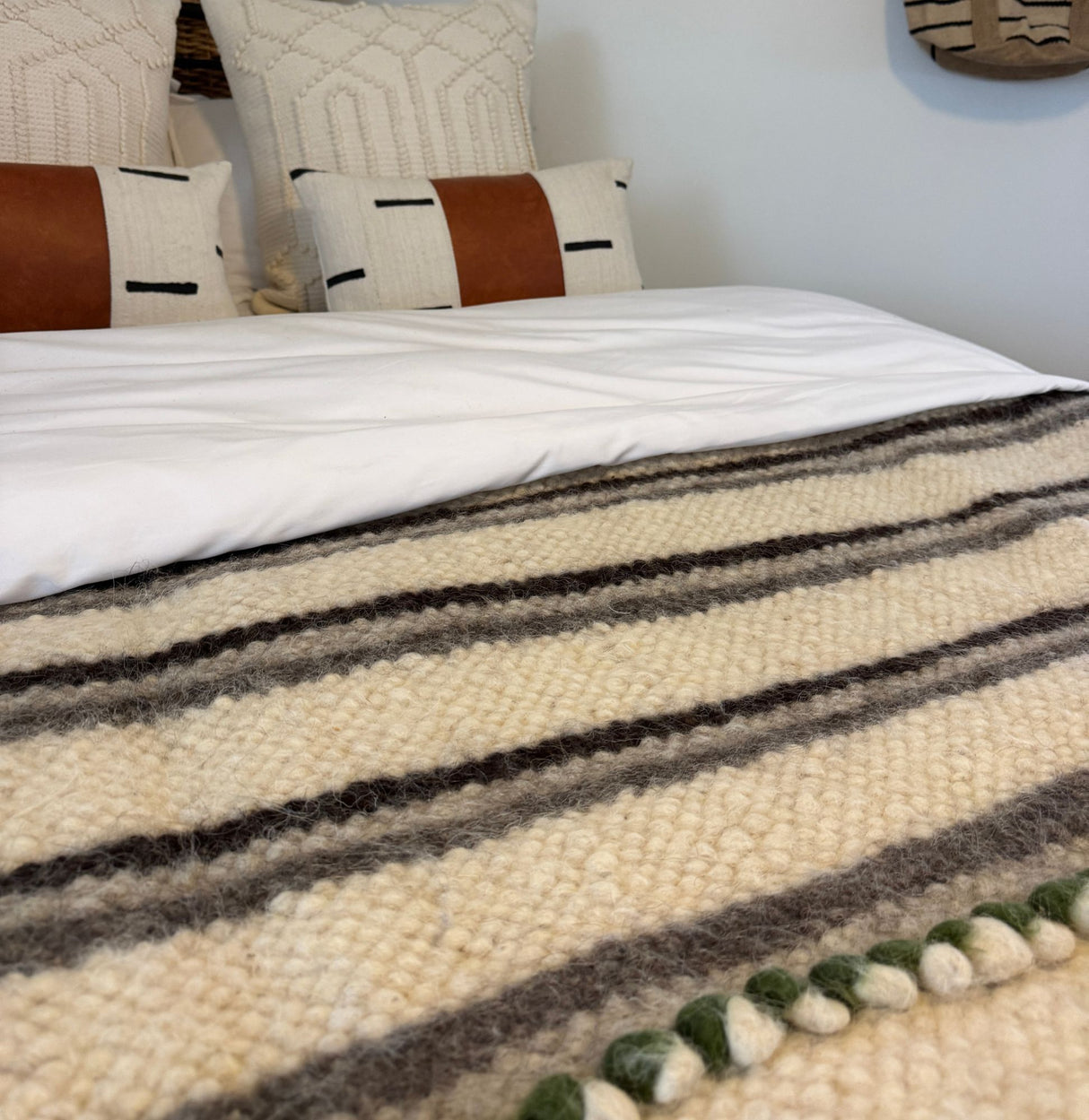 Close-up of a bed with the Faino (Fine) Wool Statement Bed Blanket and pillows, featuring brown and black accents against a white wall.
