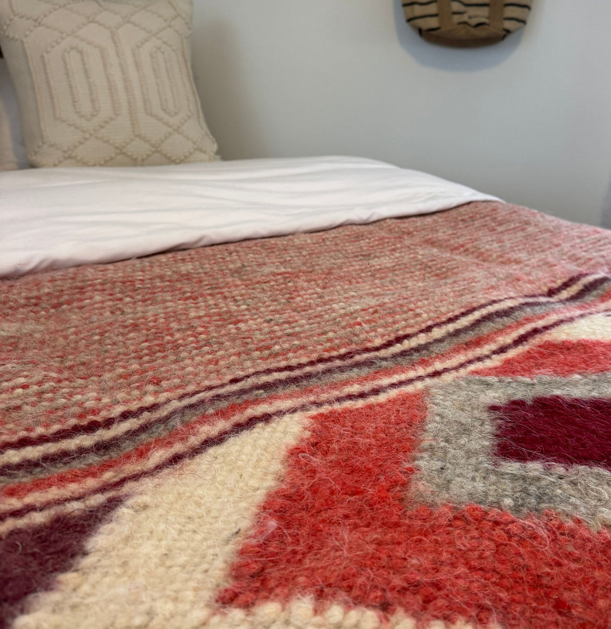 Close-up of the Tkach (Weaver) Wool Statement Bed Blanket, featuring textured red, pink, and cream handwoven wool, displayed on a neatly made bed with pillows.