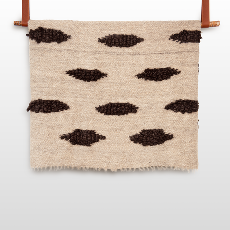 The Khmara (Cloud) Wool Statement Bed Blanket features beige woven textile with black textured ovals, hung on a wooden rod by brown leather straps—a striking accent for any modern space.