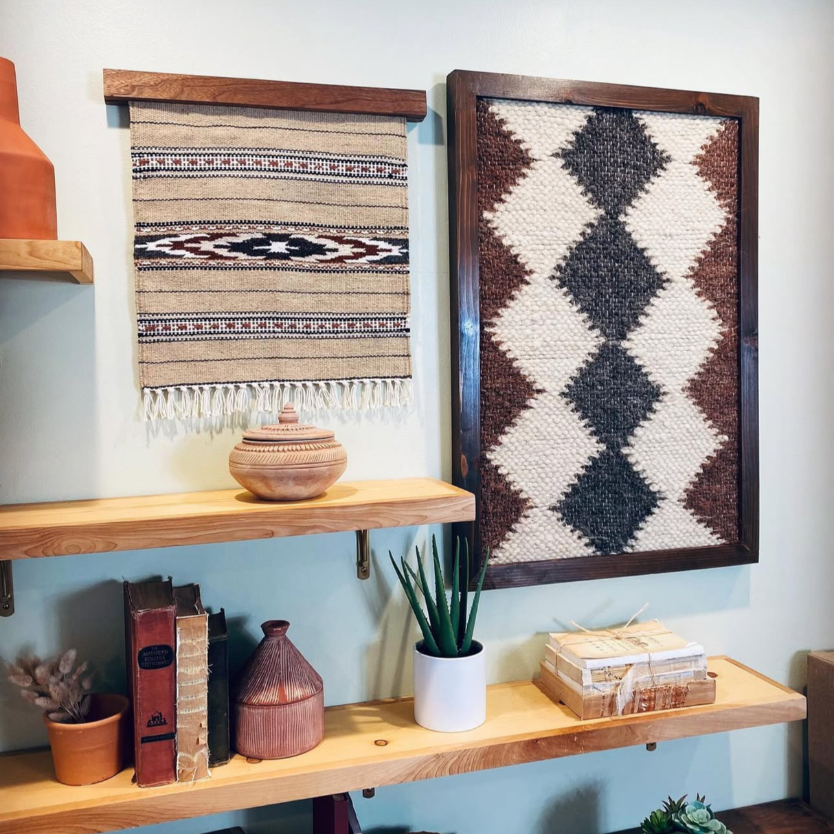 Shelves with pottery, books, plants, and two eco-friendly Dobre (Good) Wool Framed Wall Art pieces featuring geometric patterns on a light wall.