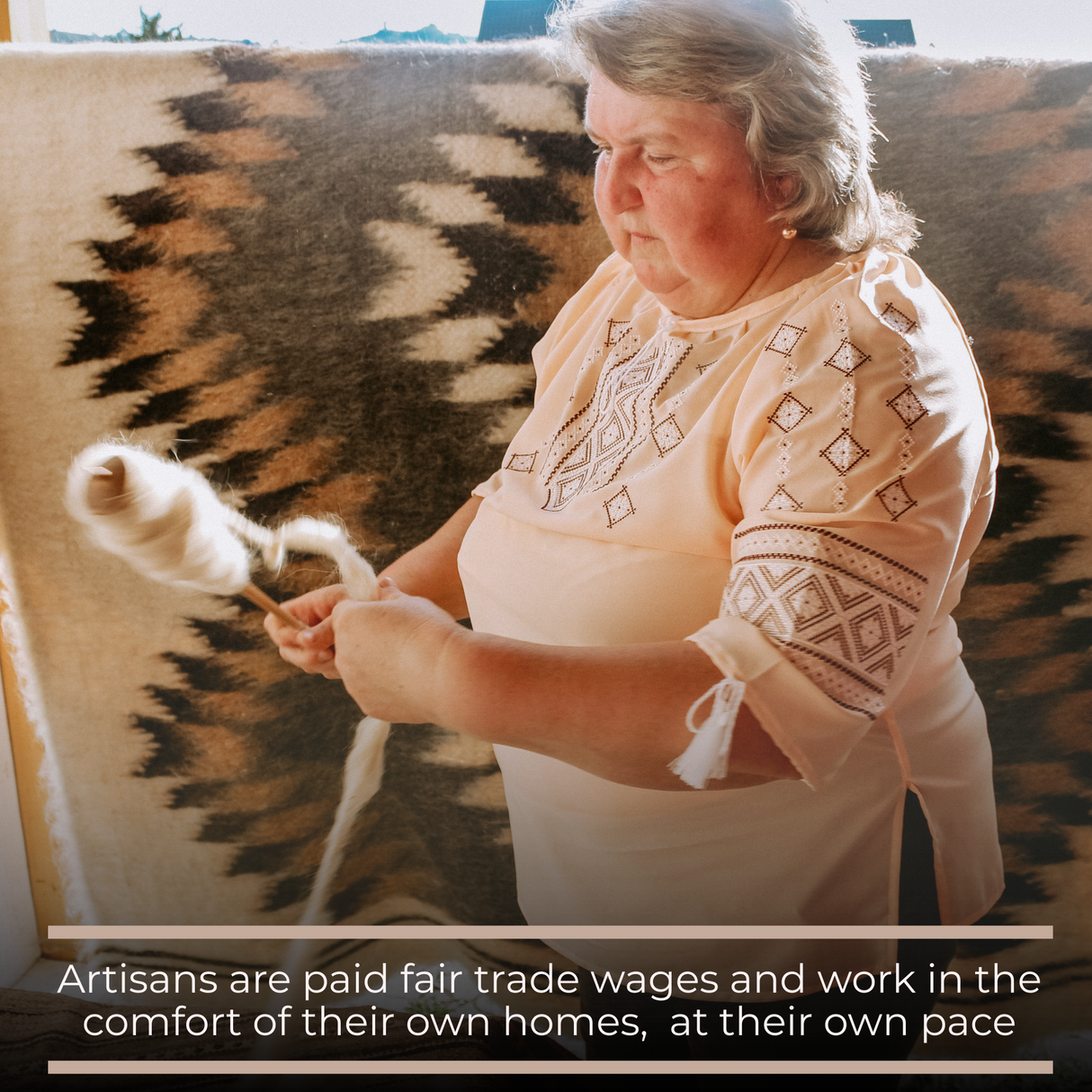 An older woman hand-spins 100% virgin wool in front of a patterned textile, proudly crafting each Korysno (Helpful) Wool Bed/Couch Runner from our Natural Collection, supporting fair trade artisan work.