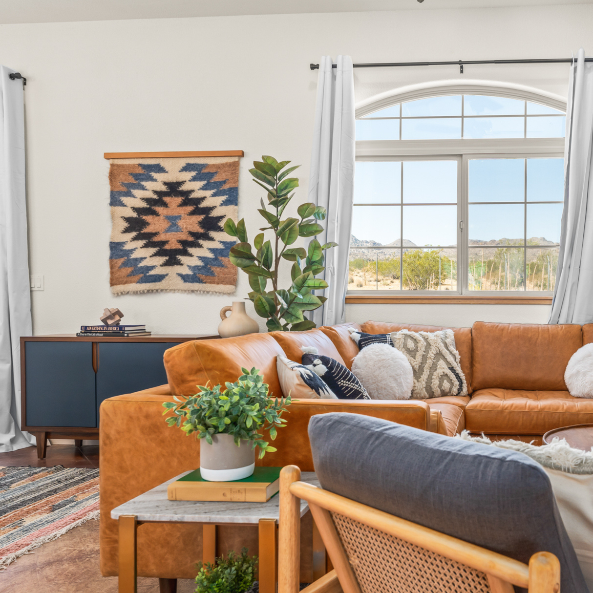 Bright living room with a tan leather sofa, plants, wall art, and a scenic window. Above the sofa, the Zirka (Star) Wool Wall Hanging, handcrafted from Carpathian sheep wool, adds a unique decorative touch.