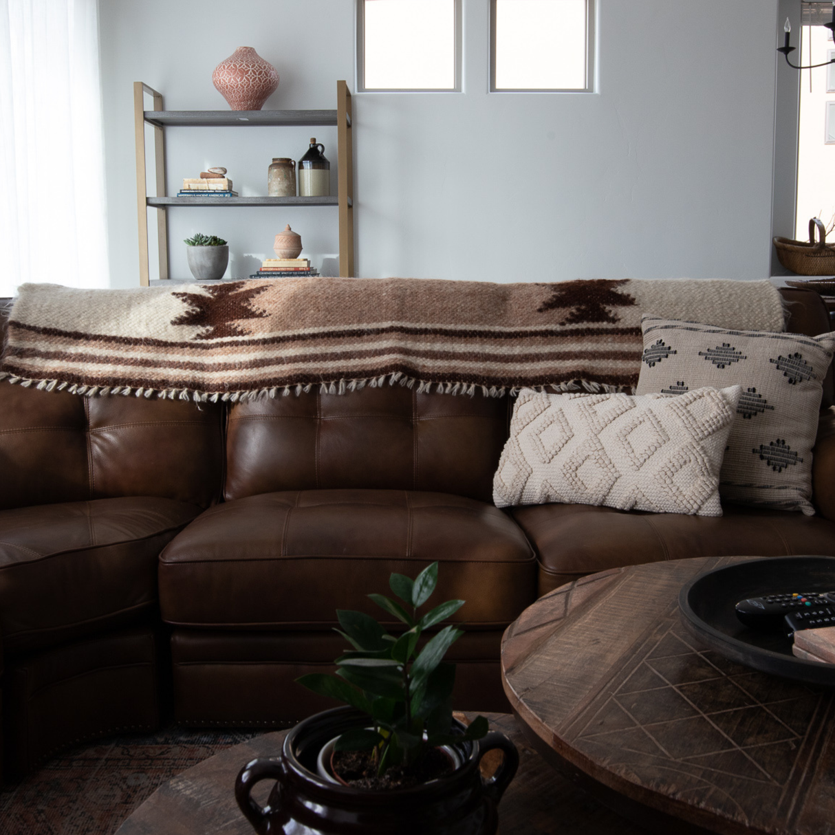 Opora (Pillar) Wool Bed/Couch Runner, handcrafted from Carpathian sheep’s wool, adds rustic charm to a brown leather sofa with patterned throws and pillows in a cozy living room with shelves and a potted plant.