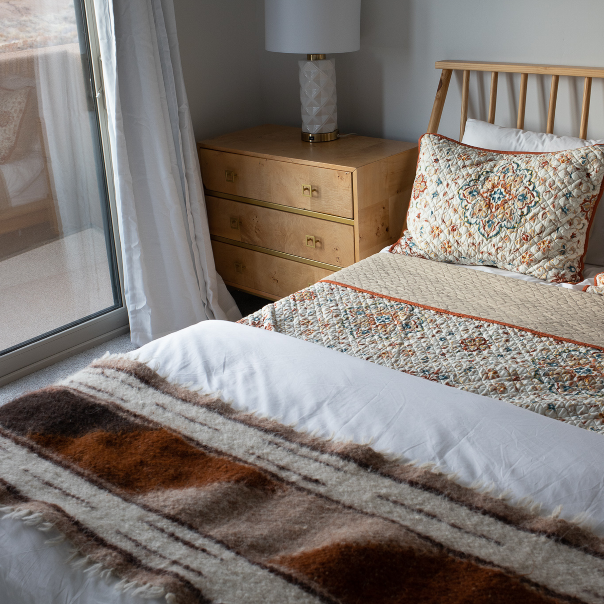 A neatly made bed sits beside a dresser and lamp near a sliding glass door, resting atop the Tak (Yes) Wool Bed / Couch Runner, which adds warmth and texture to the space.