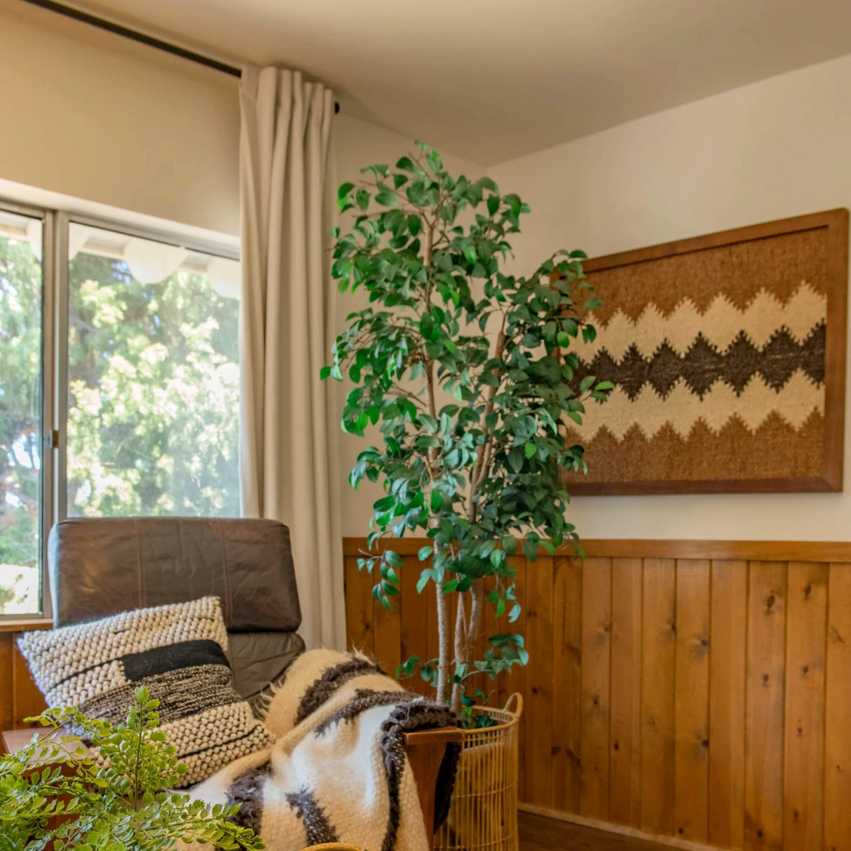 A cozy corner with a chair, pillows, blanket, potted plant, and Dobre (Good) Wool Framed Wall Art featuring organic wool decor near a sunny window.