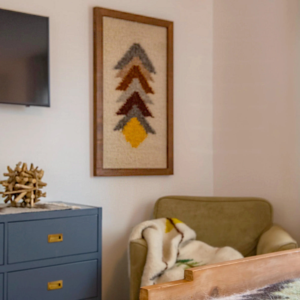 A cozy corner with an olive armchair, blanket, blue cabinet, and a Strilka (Arrow) Wool Framed Wall Art featuring geometric designs above.