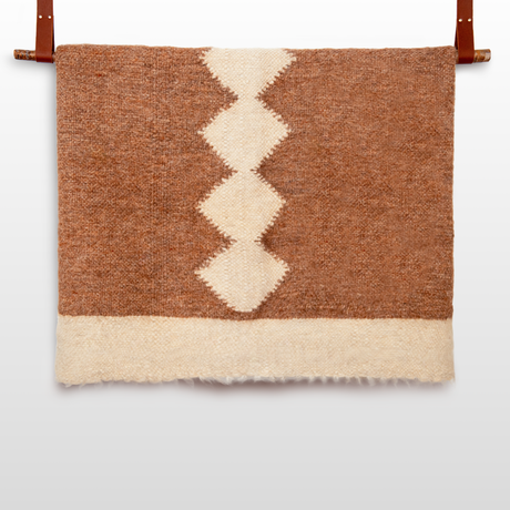 The Druh (Friend) Wool Statement Bed Blanket, in brown and cream undyed wool with a zigzag diamond pattern, hangs from two brown leather straps.