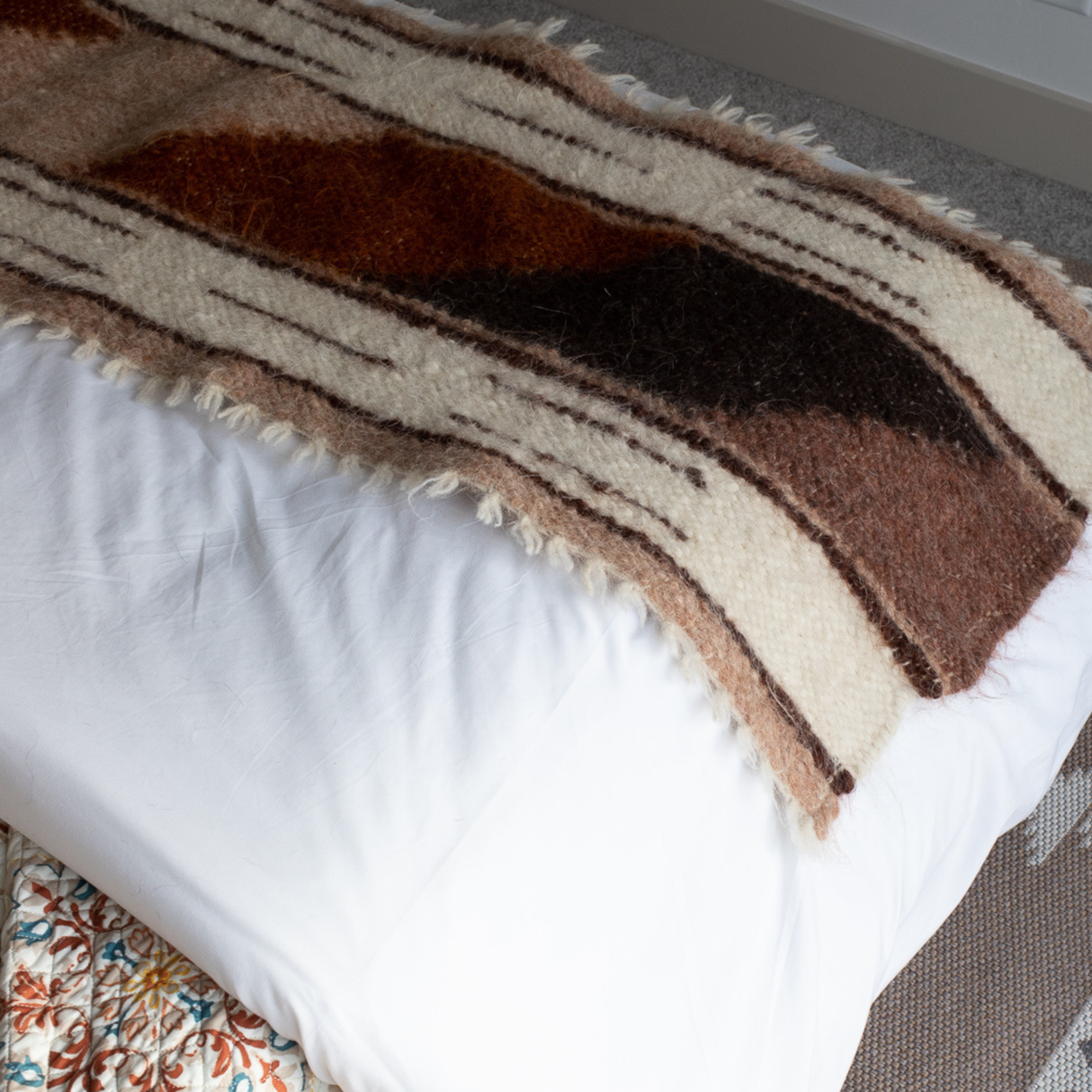 The Tak (Yes) Wool Bed / Couch Runner, featuring fuzzy brown and cream stripes, is handcrafted and lies across the foot of a neatly made white bed.