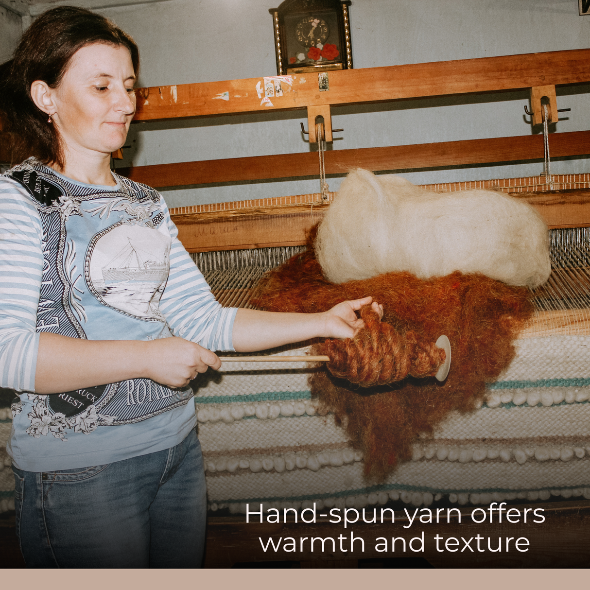 A woman spins yarn by hand in front of a loom, surrounded by wool bundles and the Faino Fine Wool Statement Throw/Baby Blanket, emphasizing its warmth and soft texture.
