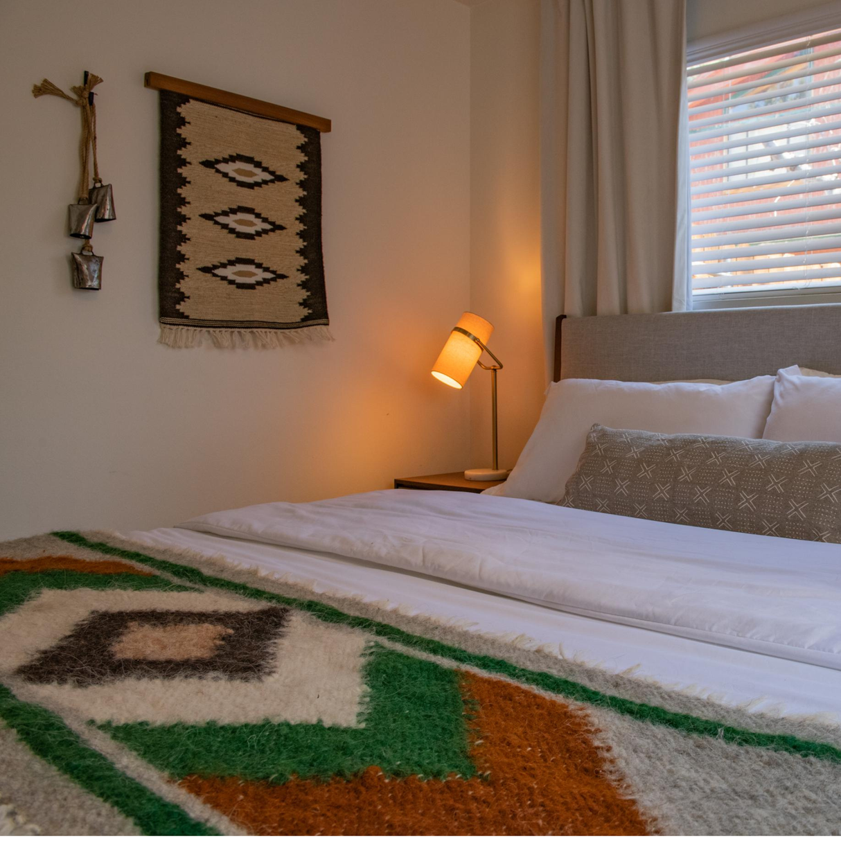 A cozy bedroom with a patterned blanket, wall art, a lit lamp, and pillows on a neatly made bed features the Heredij (Heritage) Wool Rug / Wall Hanging, crafted from naturally dyed wool.