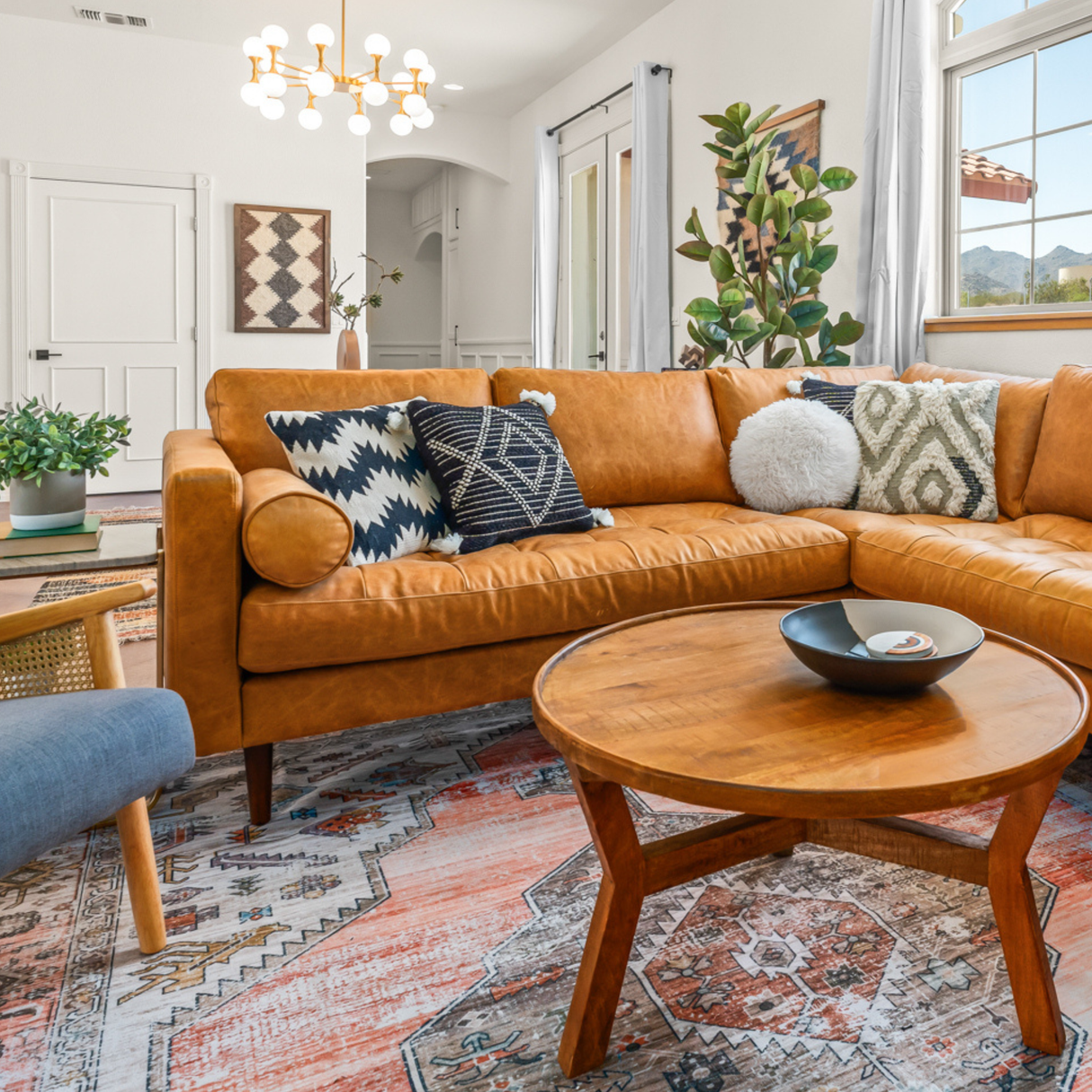 A cozy living room with a tan leather sofa, patterned pillows, a round wooden table, leafy plants, and artisanal decor featuring the Dobre (Good) Wool Framed Wall Art.