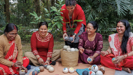 Five women in colorful clothes sit outdoors, smiling and crafting with felt balls around a basket.