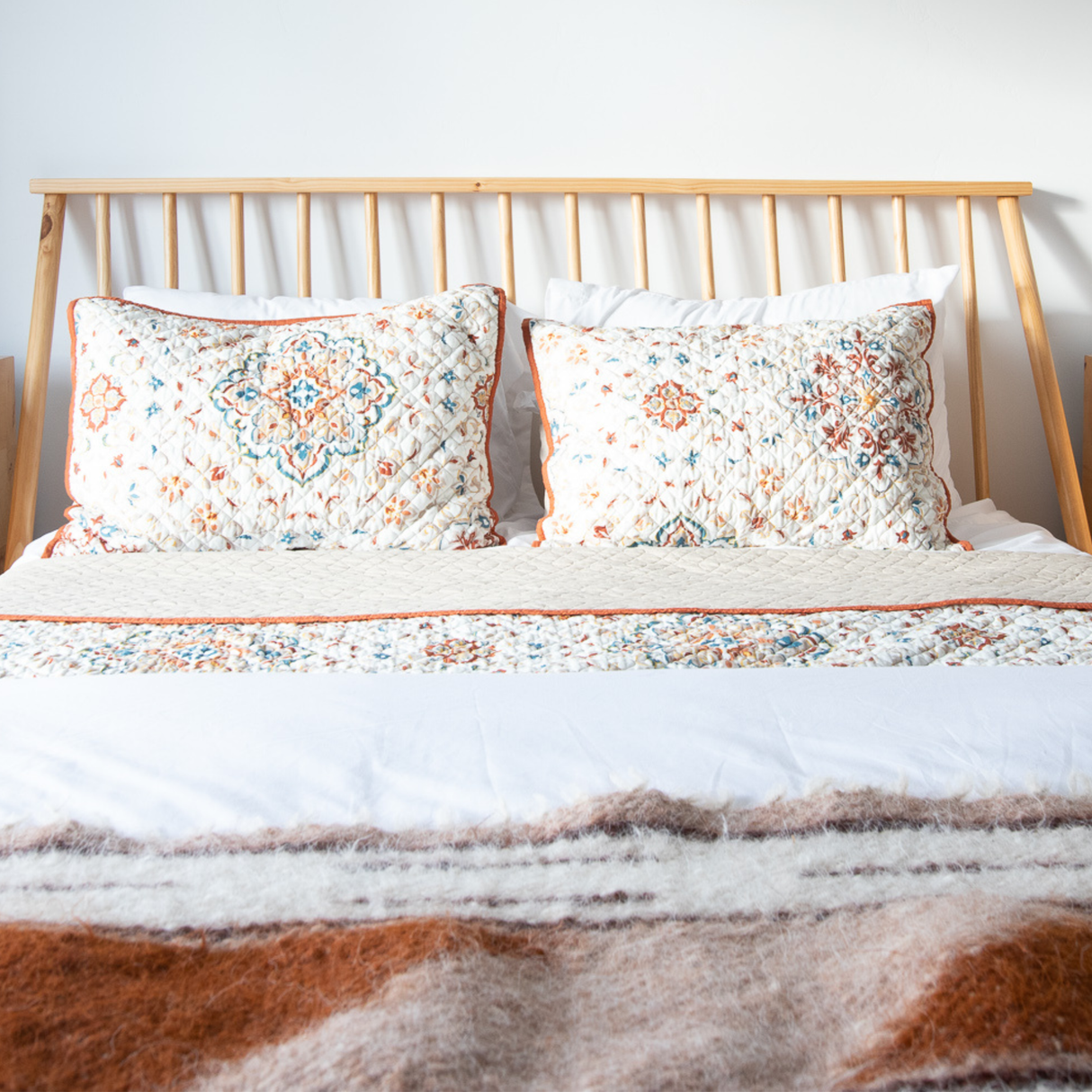 A neatly made bed with patterned pillows and a wooden headboard, covered by a brown and white blanket, sits beside the Tak (Yes) Wool Bed / Couch Runner, bringing warmth and texture to the room.