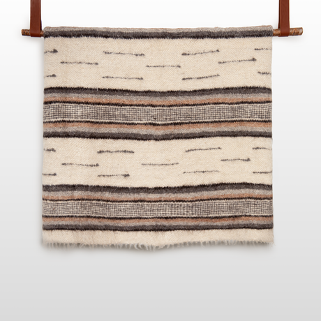 The Gratsia (Grace) Wool Statement Bed Blanket in cream and brown, with horizontal stripes and textured patterns, hangs on a wooden rod—a cozy, handcrafted accent for any space.