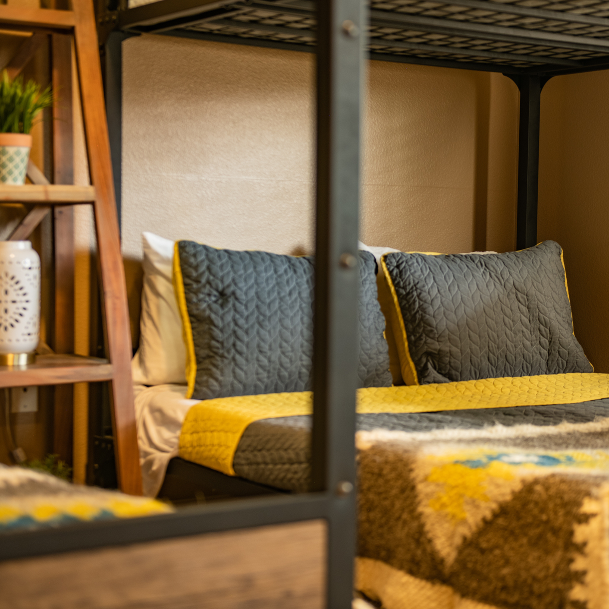 A bunk bed with gray and yellow bedding is accented by the Organic 100% Sheep Wool Bed Runner—Handmade Ukrainian Couch Cover or Bedspread from the Carpathian Mountain Collection, next to a wooden shelf with plants and decor.