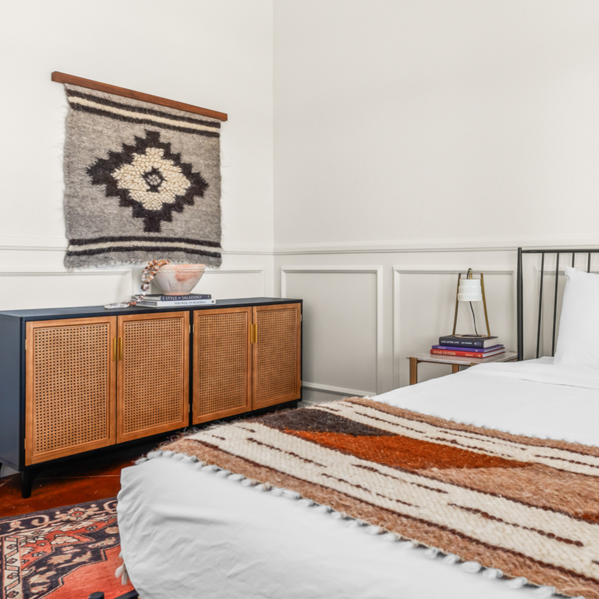 A cozy bedroom features the Tak (Yes) Wool Bed / Couch Runner, a patterned rug, woven wall art, a cabinet, and a neatly made bed with a striped blanket.