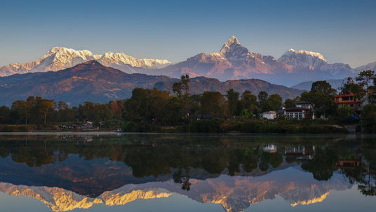 Snowy Himalayan mountains reflected in a calm lake, with trees and houses in the foreground at sunrise.