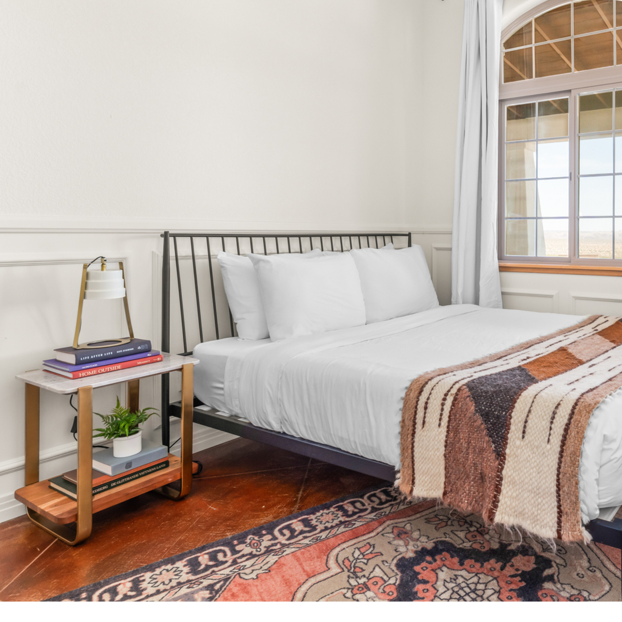 A neatly made bed with white linens sits by a nightstand topped with books, a lamp, and a small plant on the Tak (Yes) Wool Bed / Couch Runner, adding warmth and texture to the serene space.
