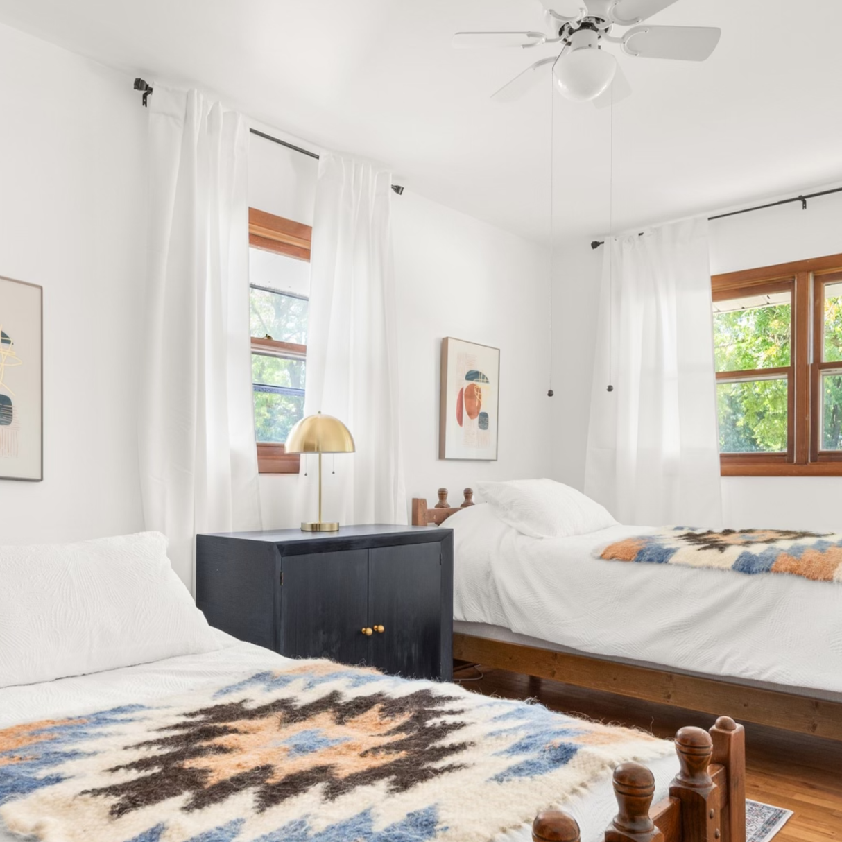 Bright bedroom with two twin beds, white bedding, and large windows with white curtains. The Zirka (Star) Wool Statement Throw adds a charming touch, while patterned blankets complete the inviting look.