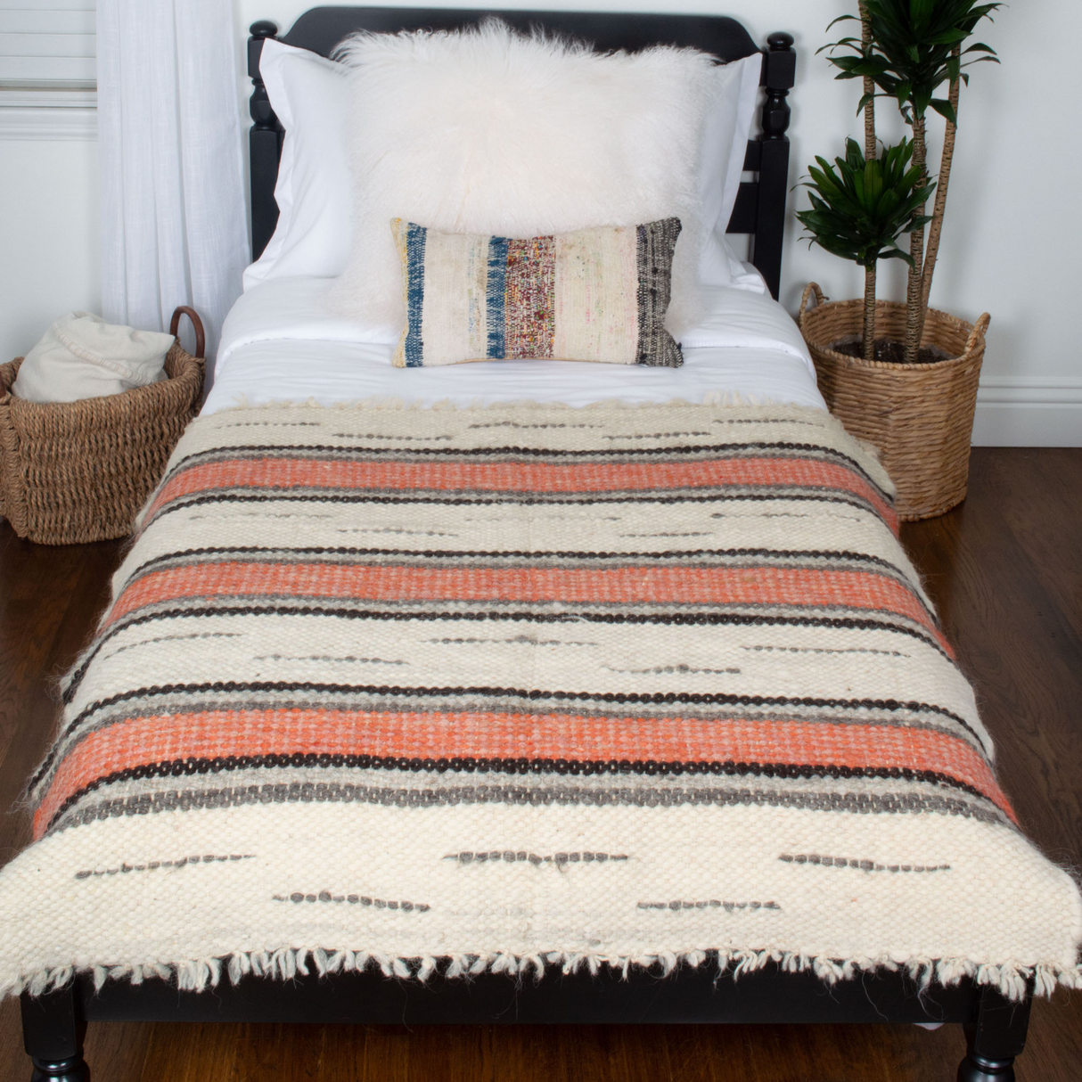 A neatly made bed features the Chudovo (Well) Wool Statement Throw/Baby Blanket, handcrafted from striped Carpathian sheep wool, accented by decorative pillows and potted plants on a wooden floor.