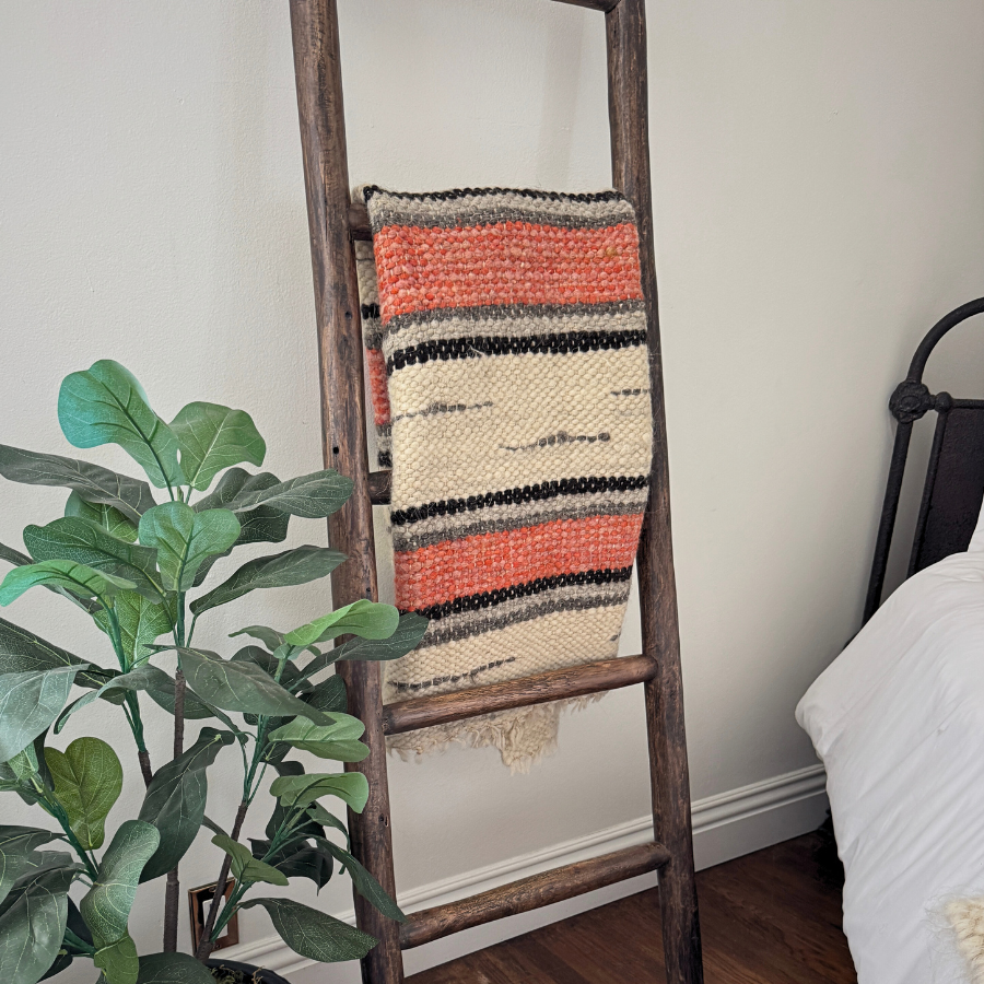 The Chudovo (Well) Wool Statement Throw / Baby Blanket, handcrafted from Carpathian sheep wool, hangs on a wooden ladder beside a potted plant and bed in a cozy room.
