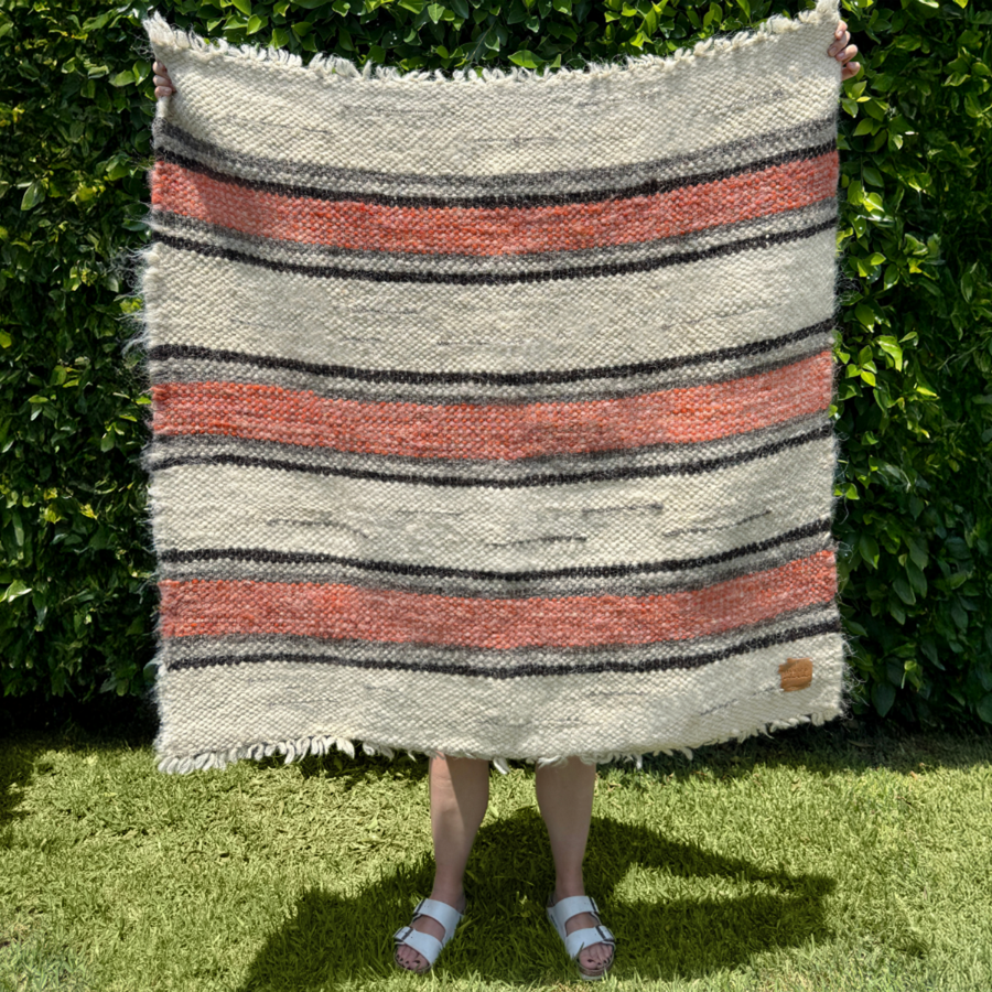 A person stands on grass before a leafy hedge, holding the Chudovo (Well) Wool Statement Throw / Baby Blanket—handcrafted from Carpathian sheep wool, featuring bold red and black stripes.