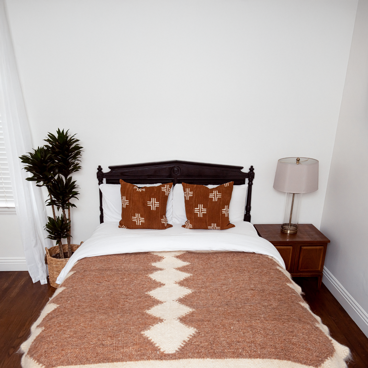A neatly made bed features brown pillows, the Druh (Friend) Wool Statement Bed Blanket, a potted plant, and a lamp on a wooden nightstand.