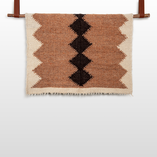 The Druh (Friend) Wool Statement Throw / Baby Blanket features brown woven textile with a black zigzag pattern and cream-colored edges, crafted from undyed wool and displayed hanging from a rod.