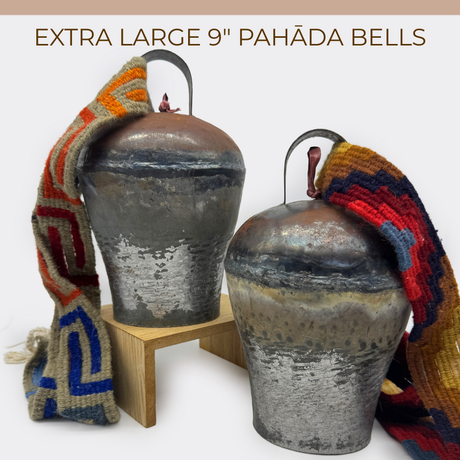 Two Vintage Handmade Pahāḍa Yak, Goat & Sheep's Bells from the Himalayan Mountains of Nepal and Tibet, each 9", are displayed with colorful woven straps on and beside a wooden stand. Perfect for Christmas ornaments, wreaths, or sound baths.