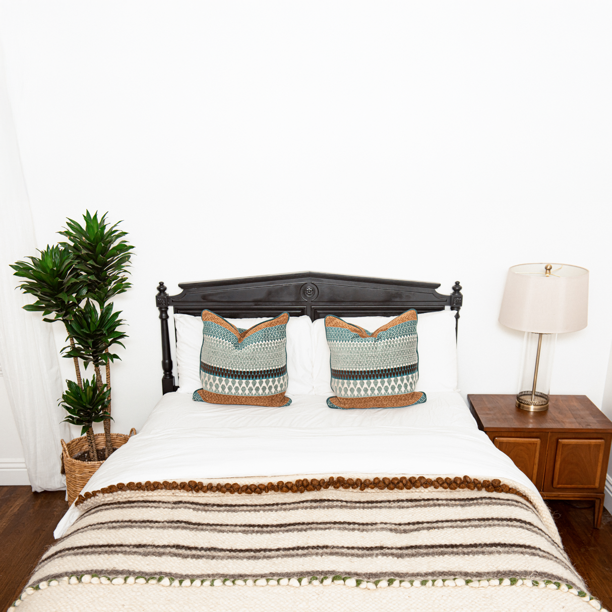 A neatly made bed with patterned pillows, the Faino (Fine) Wool Statement Bed Blanket draped across, a bedside lamp, and a leafy potted plant.